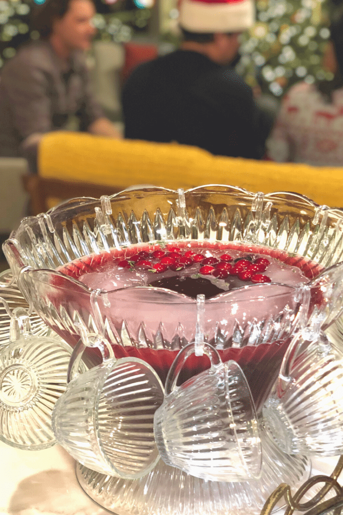 A big crystal bowl of crimson punch with glasses and a ladle with a holiday party in the background