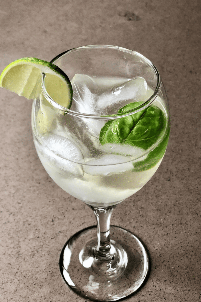 A clear, golden cocktail in a wine glass over ice with a lime and basil garnish