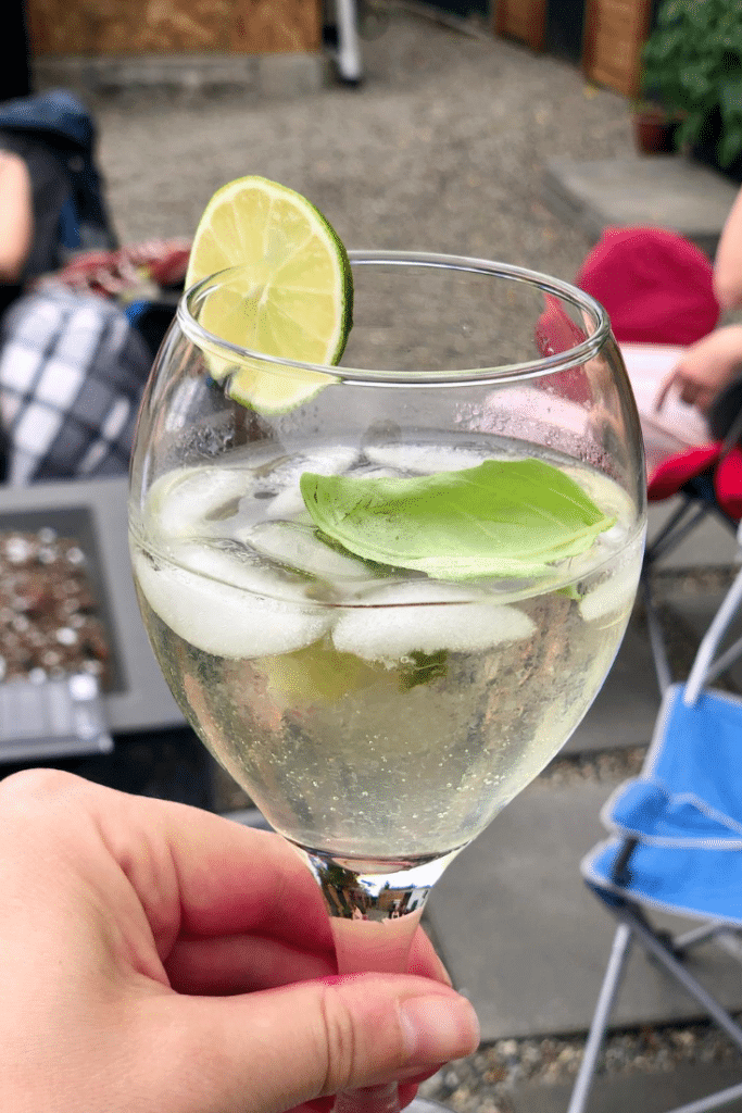 A clear, golden cocktail in a wine glass over ice with a lime and basil garnish