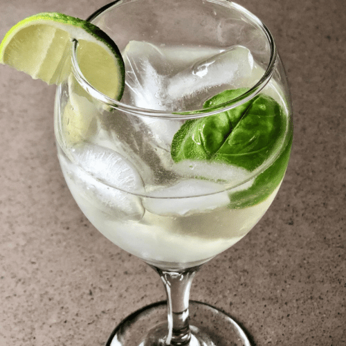 A clear, golden cocktail in a wine glass over ice with a lime and basil garnish