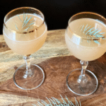 a lightly golden colored cocktail garnished with pine in a gold-rimmed glass